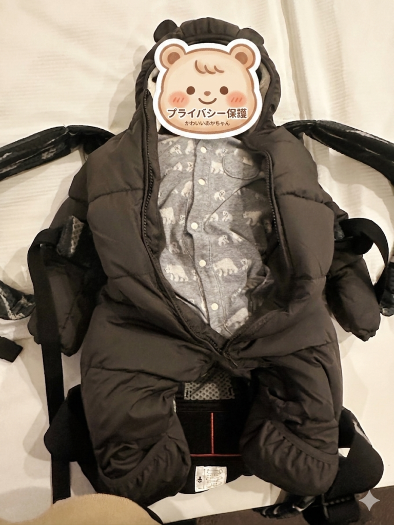 全身ダウンスーツを着た赤ちゃんが、抱っこ紐の上で寝ている正面からの写真。